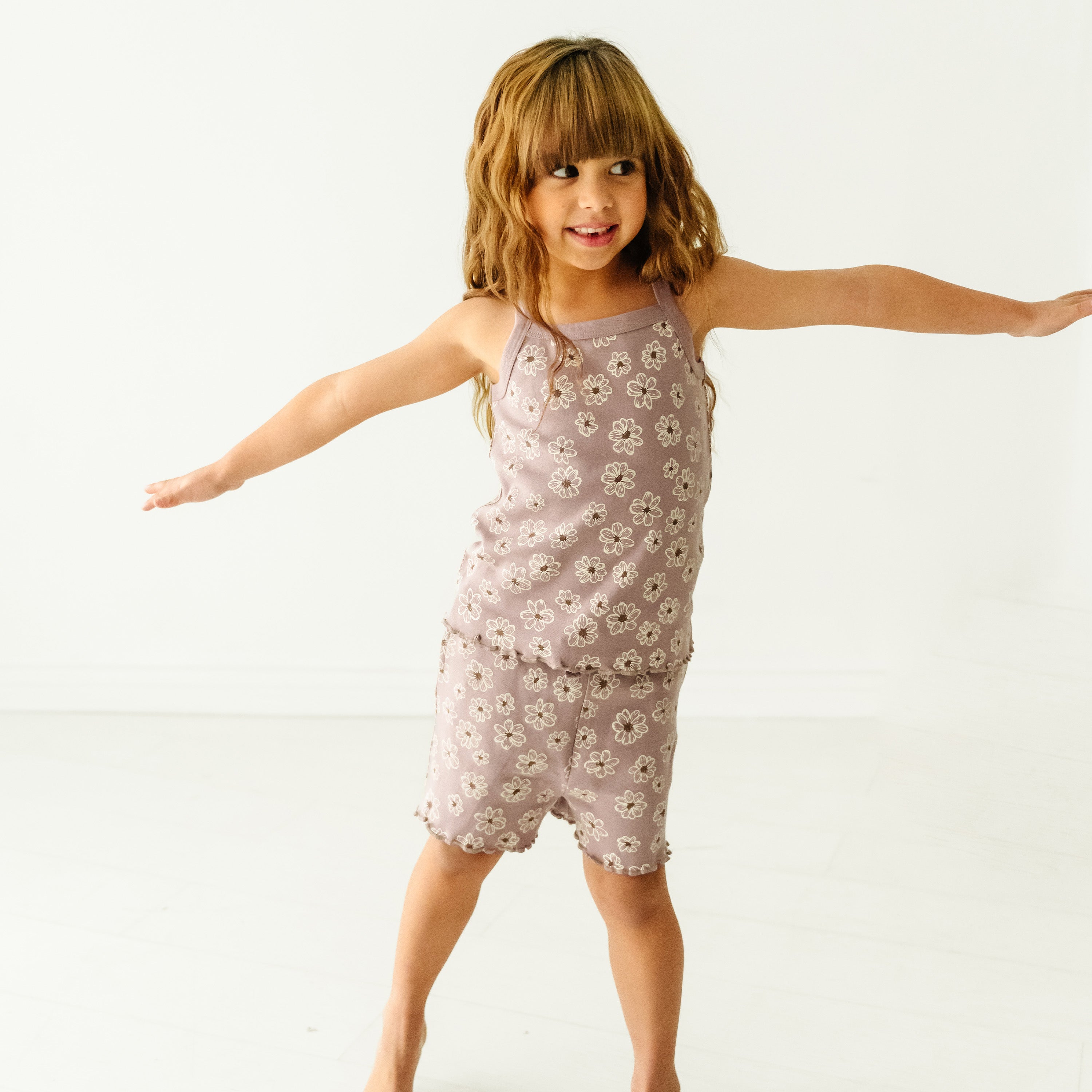 A young girl with bangs, wearing a Makemake Organics Organic Spaghetti Top & Shorts Set - Daisies, smiles and stretches out her arms while balancing in a playful pose against a white background.