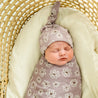 A newborn baby wrapped in a Makemake Organics Organic Swaddle Blanket & Hat - Daisies, sleeps peacefully in a woven basket on a soft, lime green pillow.