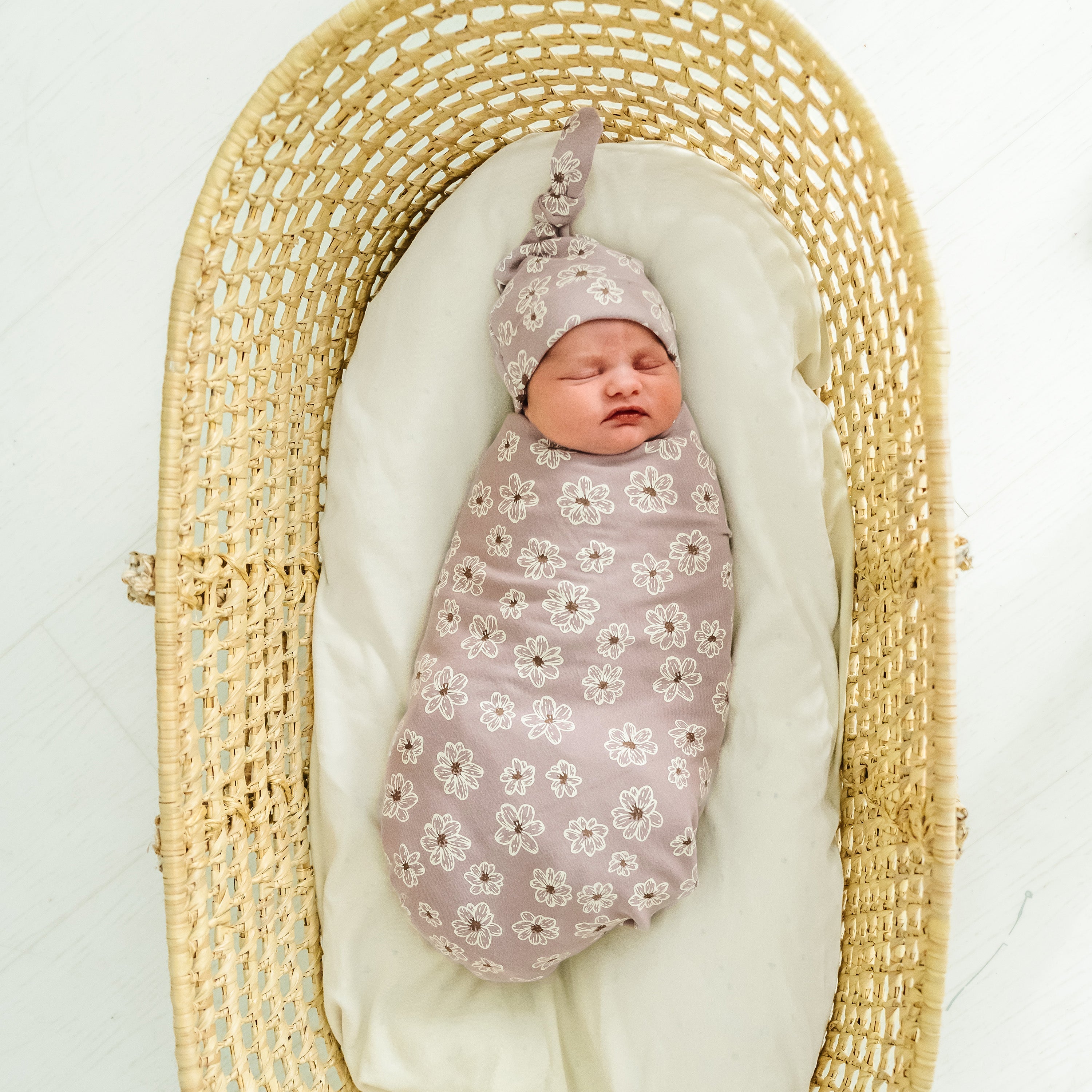 Newborn baby swaddled in a Makemake Organics Organic Swaddle Blanket & Hat in daisies pattern, lying in a woven, oval-shaped bassinet on a white background.