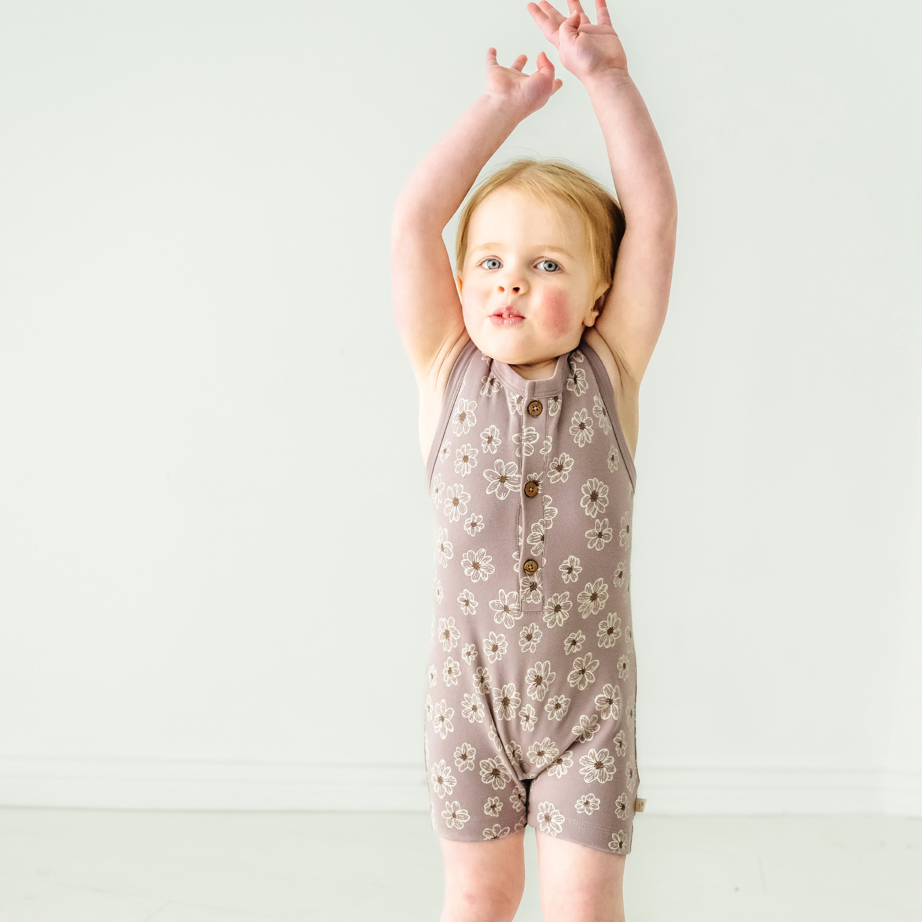 A baby in an Organic Sleeveless Short Romper - Daisies from Makemake Organics happily reaching up with both arms against a plain light background.