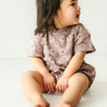 A baby wearing a Makemake Organics Organic Bubble Romper in Daisies sits on a white floor, looking to the side with a joyful expression.