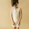 A young child wearing an Organic Tank & Shorts Set - Speckle from Organic Kids, and white sunglasses, smiling against a beige background.