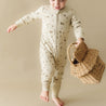 Toddler in an Organic 2-Way Zip Romper - Camplife by Organic Baby, holding a wicker basket, stepping forward on a beige background.
