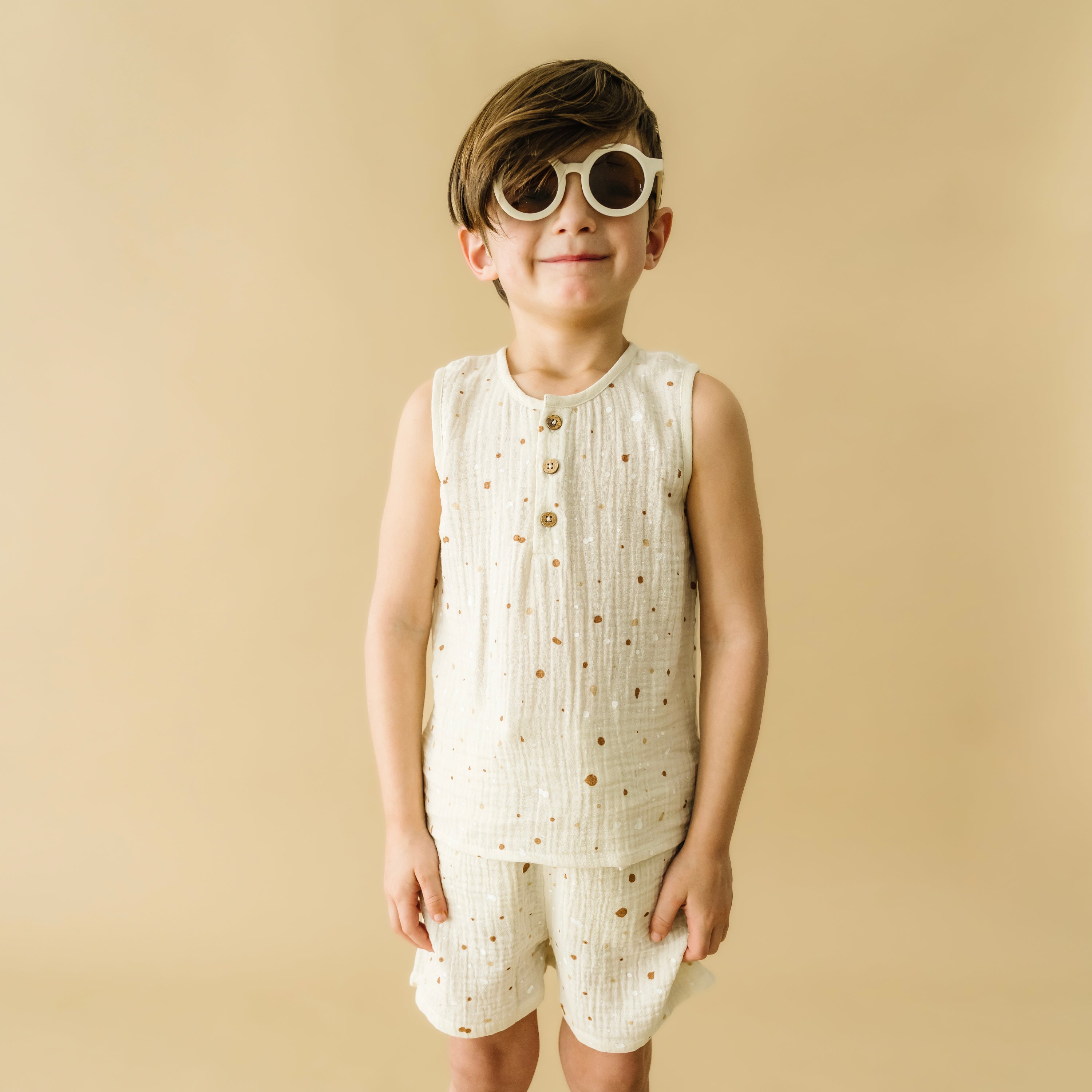 A young boy wearing a stylish white sleeveless top and shorts set from Organic Kids with matching round white sunglasses, smiling against a beige background.