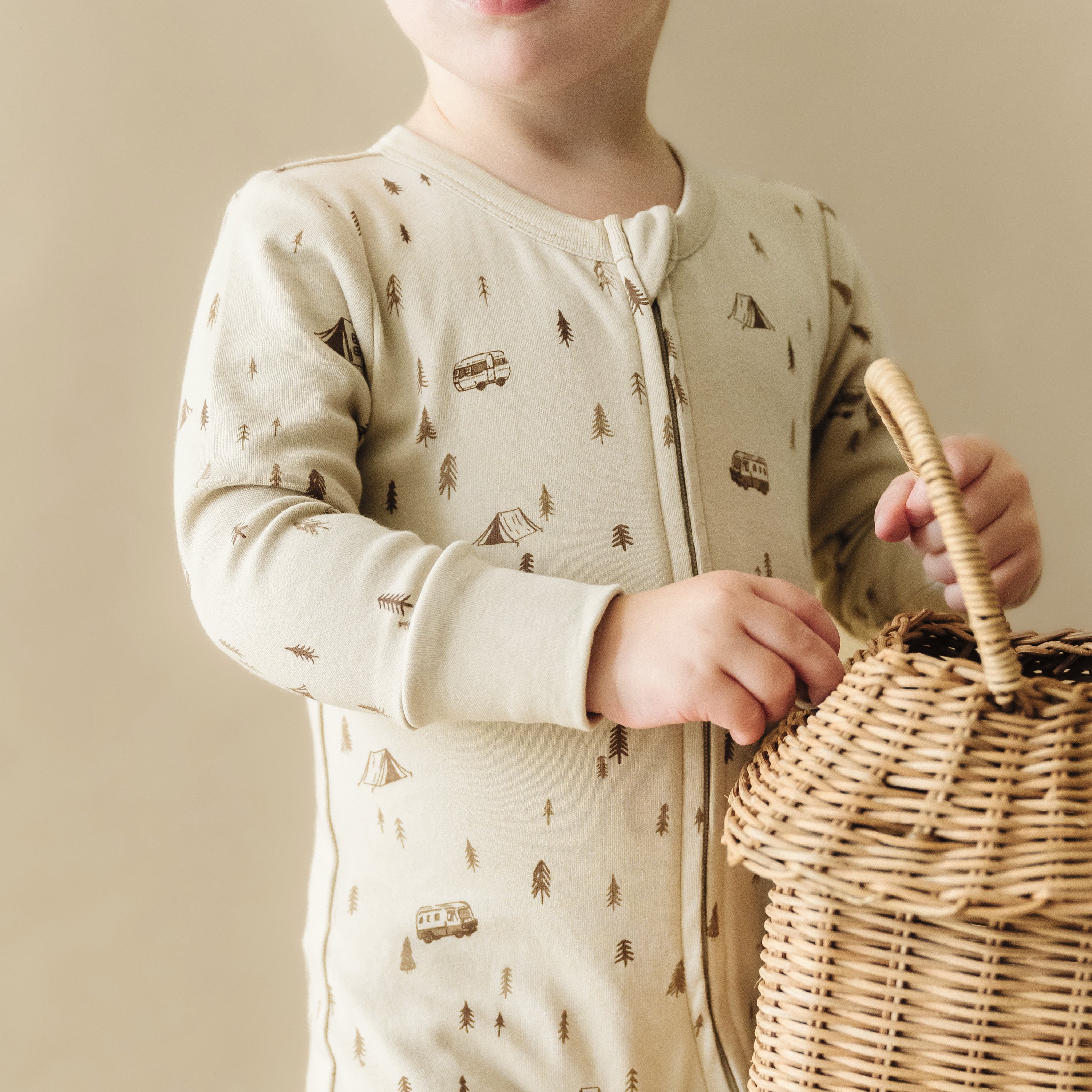 A young child in an Organic Baby Organic 2-Way Zip Romper - Camplife printed with small trees and campers holds a wicker basket, visible from the waist up against a neutral background.