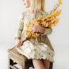 A young girl in a Organic Long Sleeve Twirl Dress - Camplife from Organic Baby sits holding a large bouquet of yellow flowers, her face not visible, against a white background.