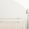 A minimalist baby nursery featuring a white crib from Makemake Organics with a Crib Fitted Sheet with Pillowcase in Polka Dots and a standing lamp in a room with light beige walls.