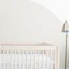 A minimalistic nursery room featuring a white crib with a Bloom crib fitted sheet and pillowcase set beside a standing lamp, set against a plain wall with a large semicircle painted on it by Makemake Organics.