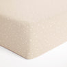 A close-up view of a beige Makemake Organics crib fitted sheet with pillowcase in a polka dot pattern, focusing on the neatly tailored corner seam and the dotted fabric texture.