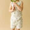 A child in an Organic Kids palm tree-patterned Organic Tee & Shorts - Tropical outfit holds a magnifying glass in front of their face, obscuring it, while holding a fern leaf over the glass. The background