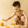 A young boy in a brown outfit from Organic Kids reaches playfully for oranges from a mesh bag on a beige background.