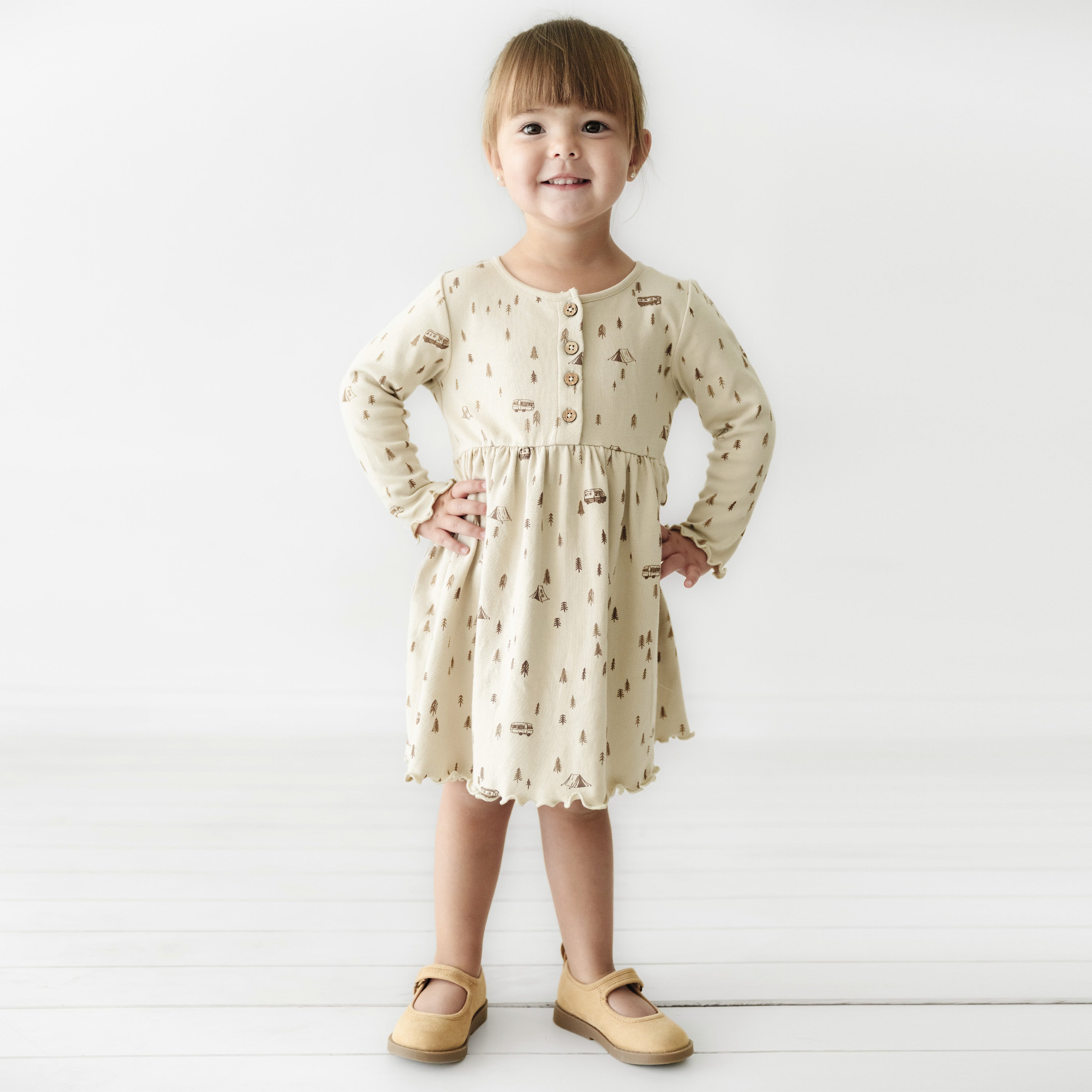 A young girl with light brown hair smiles and stands confidently, wearing an Organic Baby Camplife Long Sleeve Twirl Dress adorned with whimsical prints from Makemake Organics and brown mary jane shoes against a white background.