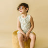 A young boy wearing Makemake Organics sunglasses and a Wild Safari Organic Kids tank top sits smiling on a wicker stool against a plain beige background.