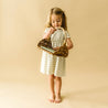 A young girl in a Organic Puff Sleeve Dress - Foam Stripes from Organic Kids is standing and looking into an empty wicker basket she is holding. The background is a plain beige color.
