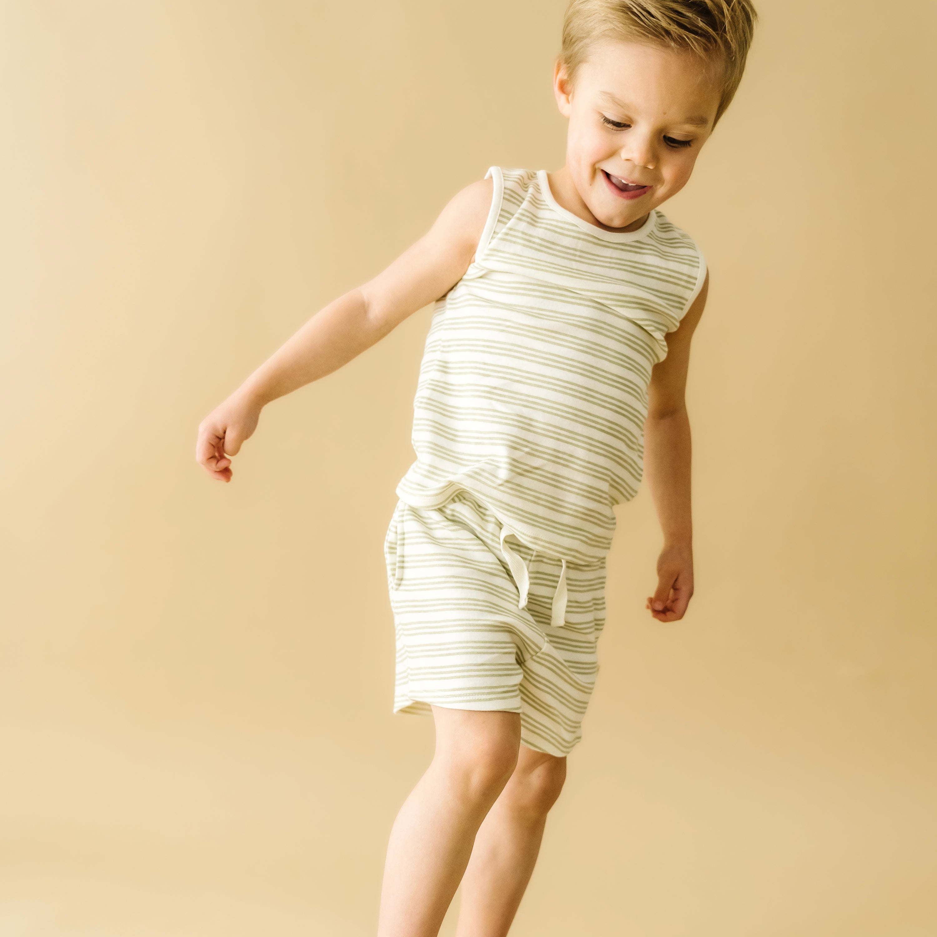 A young boy in Organic Kids Foam Stripes organic tee and shorts is playfully jumping, with a joyful expression, against a soft beige background.