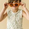 A young child playfully holding up oversized orange sunglasses to their face, wearing a sleeveless shirt by Organic Kids patterned with tropical motifs on a beige background.
