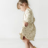 A young girl in an Organic Baby Camplife beige dress with feather prints twirls playfully in a bright room, her hair tied back, and her feet bare, evoking a sense of