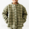A young child in a green checkered jacket from Organic Kids smiles, with only the lower half of their face visible. The background is a plain light color, emphasizing the jacket's pattern.