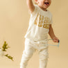A joyful toddler wearing a white Organic Tee & Pants Set - Wild Child from Organic Kids and white pants with giraffe patterns, raising one arm and holding glasses in the other, on a beige