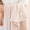 A close-up of a baby wrapped in a Chunky Knit Throw Blanket - Nora Shell with a visible brand tag that reads "Makemake Organics," held by an adult in a white blouse.