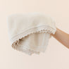 A person's arm holding an Organic Cotton Scalloped Baby Blanket in Vanilla Natural by Makemake Organics, against a light pink background.