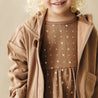 A young child with curly blonde hair smiling, wearing an Organic Baby brown jacket over a star-patterned dress, cropped view showing from chest up.