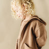 A young child with curly blonde hair, dressed in a brown Organic Hooded Jacket - Cocoa from Organic Baby, looking downward with a soft, neutral background.