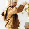 A young child with blonde hair in an Organic Baby mustard hoodie reaches playfully towards a bouquet of light pink flowers on a wooden table, portraying joy and innocence.