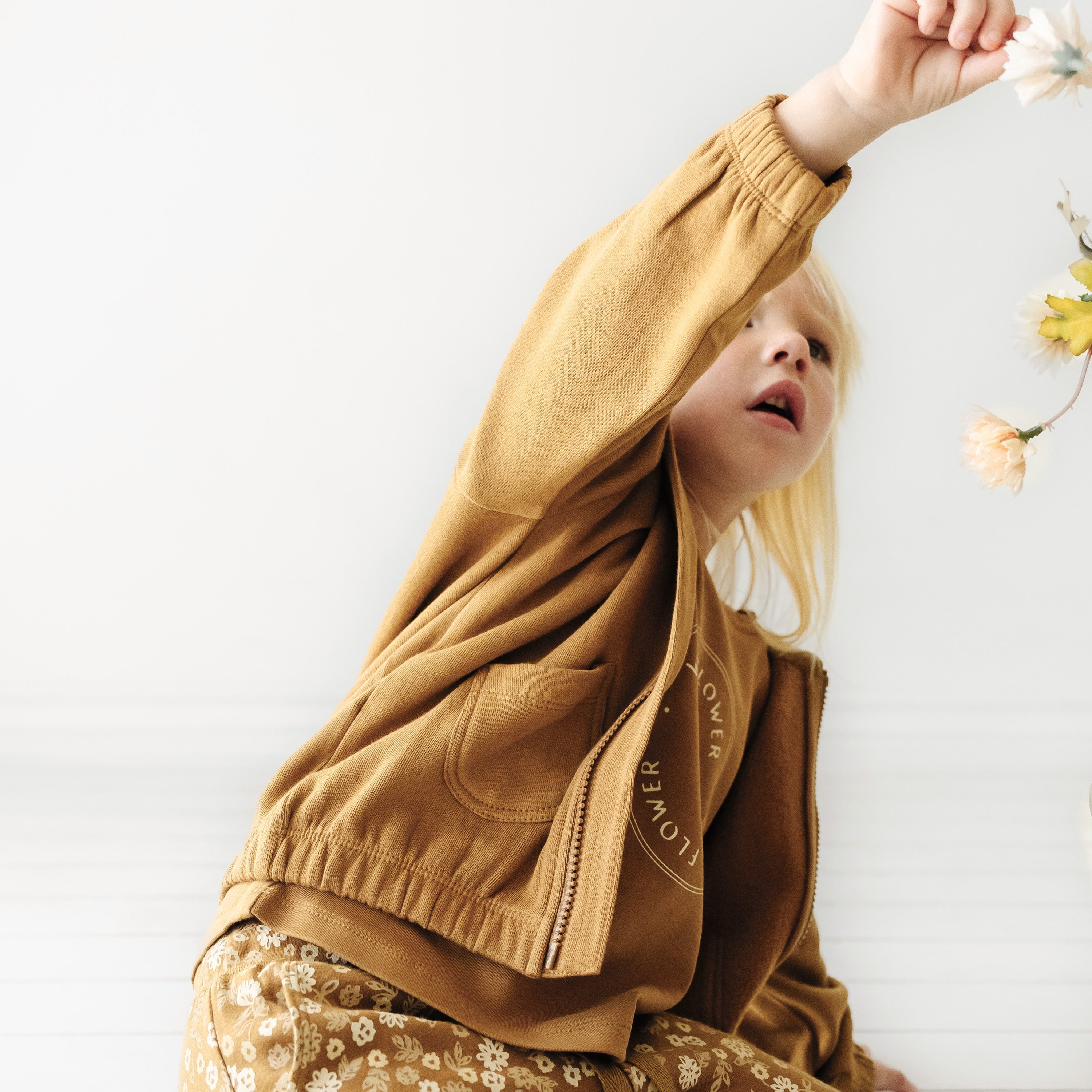 A young child with blonde hair reaching up to touch hanging flowers, wearing an Organic Baby tan Organic Hooded Jacket and patterned pants, against a white background.
