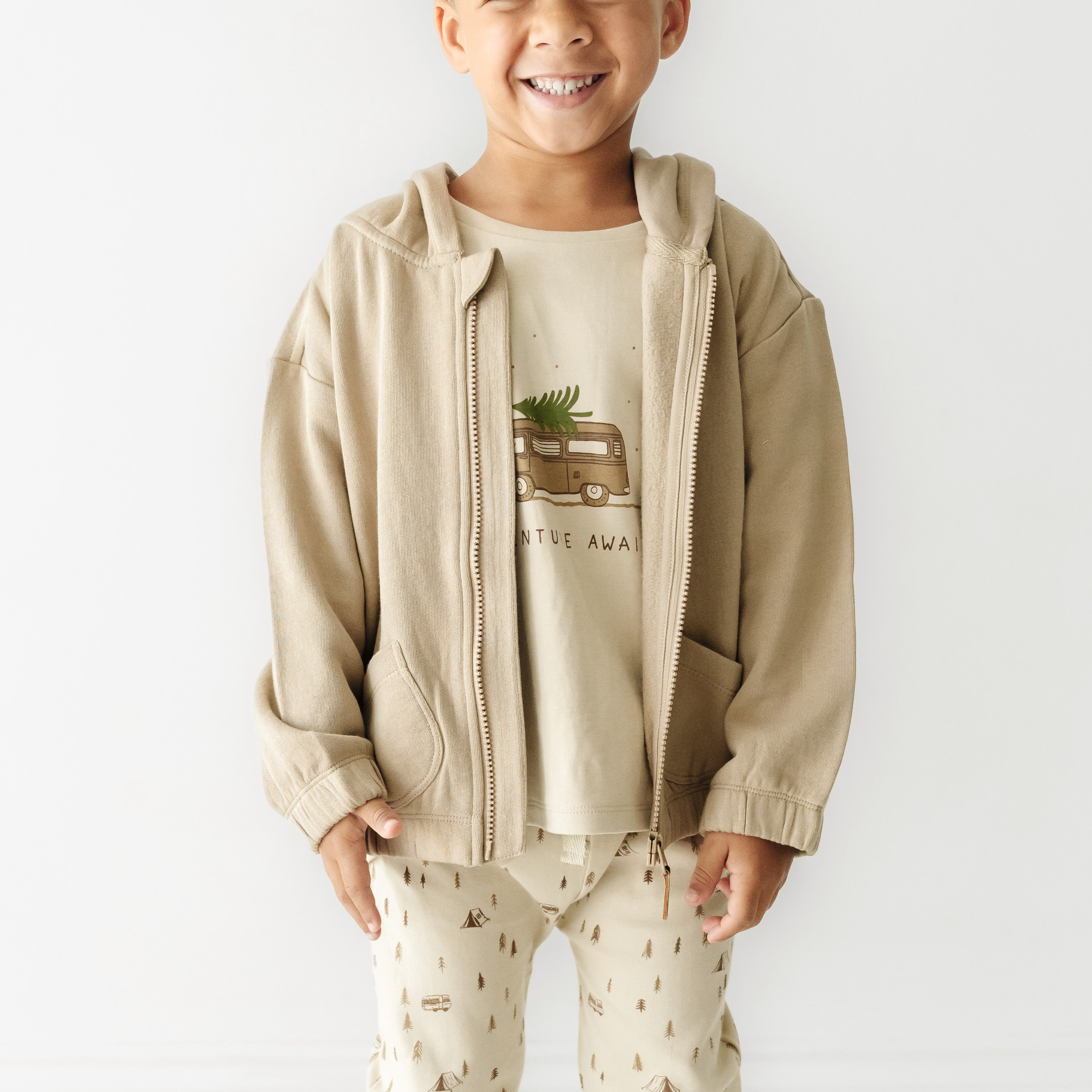A young child smiling, wearing a Organic Baby Hooded Jacket in Mocha with a graphic of a van and "adventure away" text, paired with matching pants printed with teepees and trees.