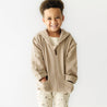 A smiling young boy in an Organic Baby mocha hooded jacket and patterned pants stands against a white background, hands tucked into his pockets.