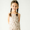 A young girl with braided hair and a sleeveless patterned dress standing against a plain background smiles gently at the camera wearing the Organic Spaghetti Top & Shorts Set in Seashells by Makemake Organics.