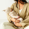 A mother lovingly holds her toddler, who is wearing a Makemake Organics Organic Kimono Knotted Sleep Gown - Seashells, in a cozy, warmly lit setting. The mother is dressed in a green cardigan and cream pants, exuding a calm and content demeanor.
