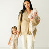 A smiling woman in a beige sweater stands holding a toddler in a matching outfit and clasps hands with a young girl in a floral dress, against a plain white background wearing the Makemake Organics Organic Kimono Knotted Sleep Gown - Seashells.