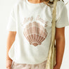 Close-up of a toddler wearing a white Boxy Tee and Skort Set - Seashells from Makemake Organics, complemented by a beige shoulder bag and patterned shorts.