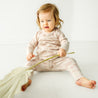 A baby in Makemake Organics' Organic Kimono Top & Pants Set -Seashells sits on a white floor, holding a large leaf, looking slightly to the side with a focused expression.