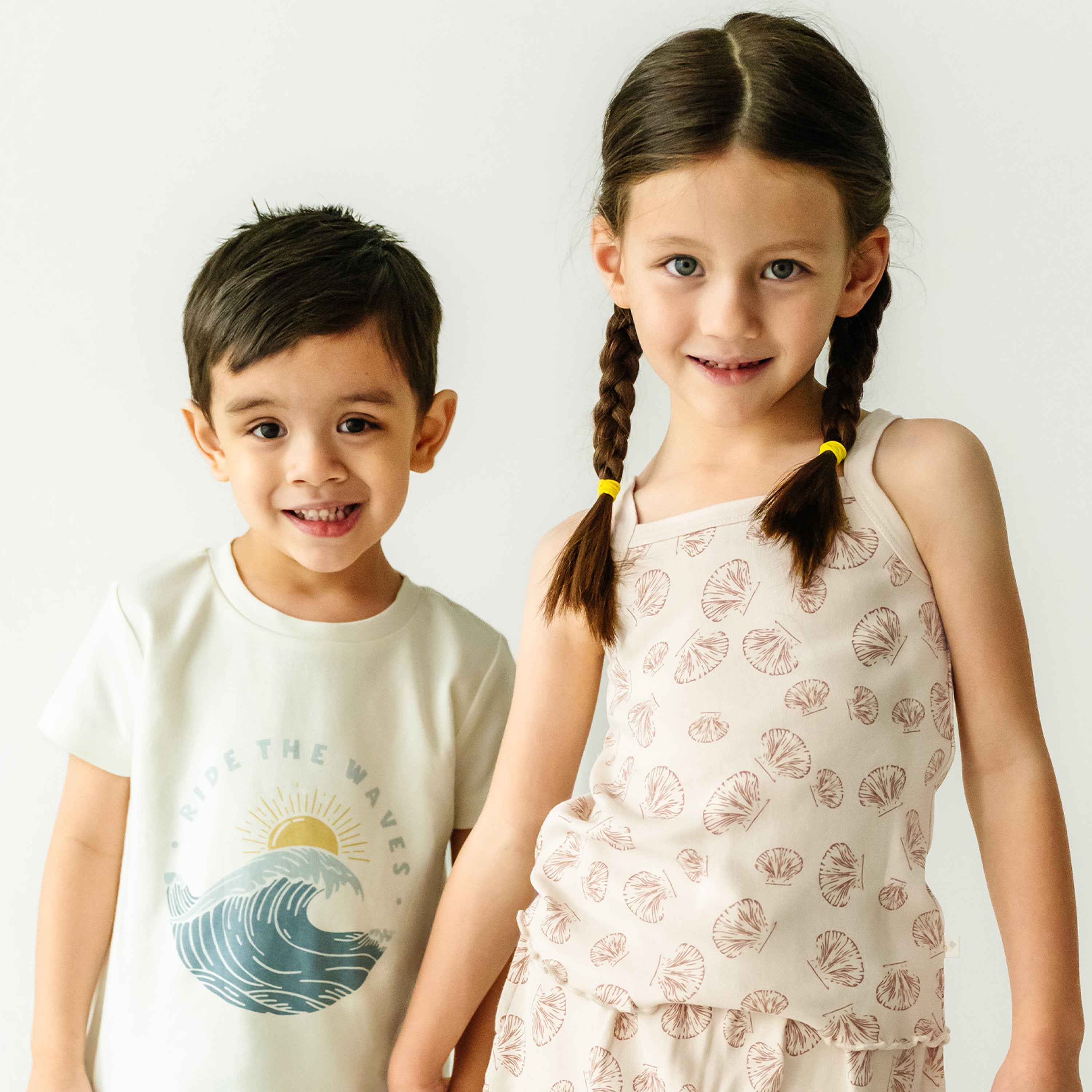 Two young children, a boy and a girl, stand smiling against a white background. the boy wears a white t-shirt with a wave graphic, and the girl wears the Organic Spaghetti Top & Shorts Set - Seashells by Makemake Organics.