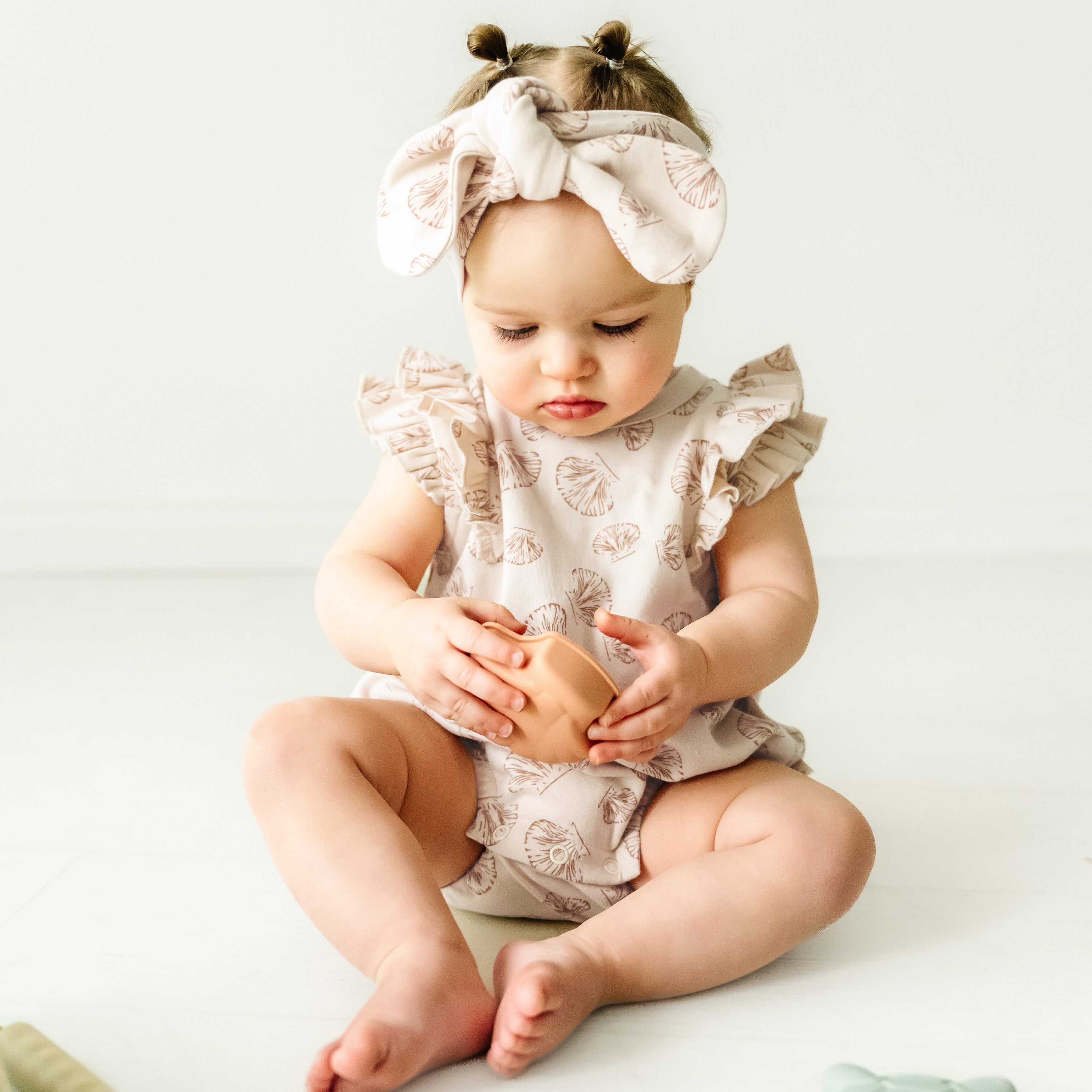 A toddler girl wearing a headband and ruffled outfit sits on the floor, examining an  Organic Flutter Bubble Onesie - Seashells in her hands, with a white background. (Makemake Organics)