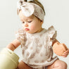A baby with a headband plays with a toy, wearing a Makemake Organics Organic Flutter Bubble Onesie - Seashells and looking curiously at a toy part in her hand.