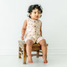 A baby with curly hair sits on a small wooden stool, wearing a Makemake Organics Organic Sleeveless Short Romper in Seashells. The child looks to the side with a curious expression against a plain, light background.