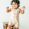 A joyful baby with curly hair, standing and smiling in a Makemake Organics Organic Sleeveless Short Romper - Seashells decorated with leaf patterns, next to a wooden stool against a plain white background.