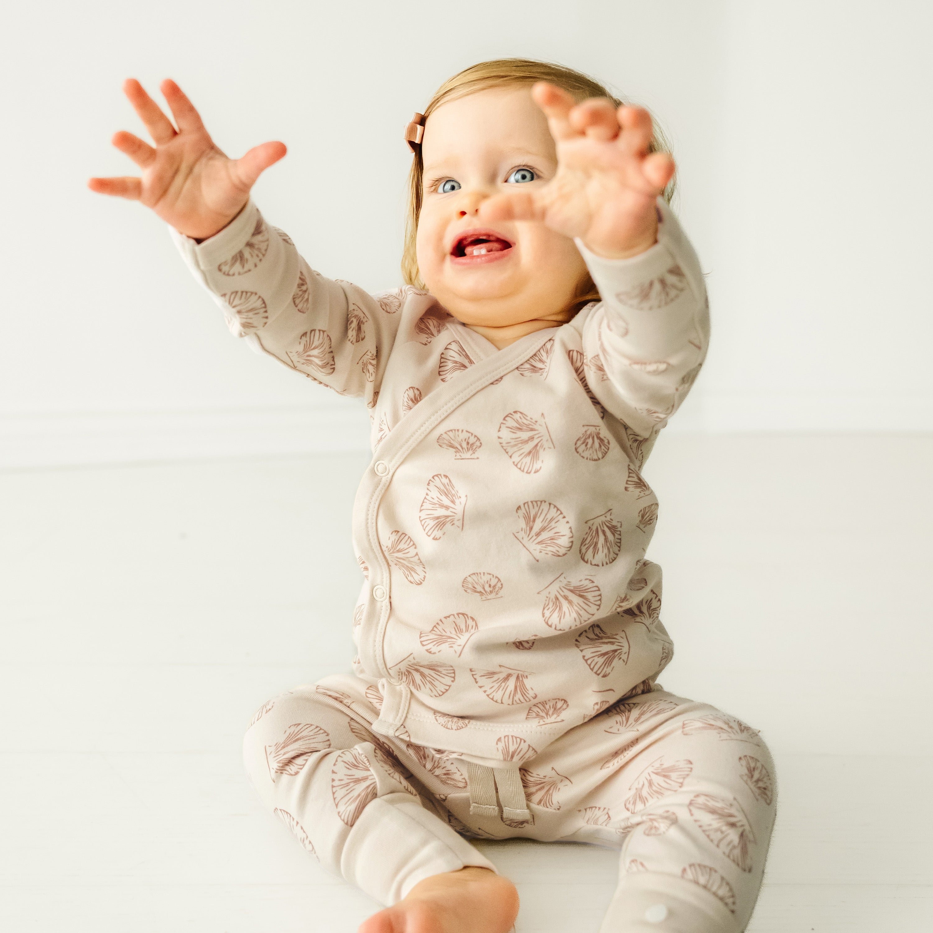 A joyful toddler in a Makemake Organics Organic Kimono Top & Pants Set - Seashells patterned with leaves reaches up with both hands, smiling widely, seated against a plain white background.