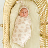 A newborn baby sleeps peacefully in a yellow Organic Swaddle Blanket & Hat with a leaf pattern, lying in a woven bassinet. The baby wears a knotted beige hat on their head.