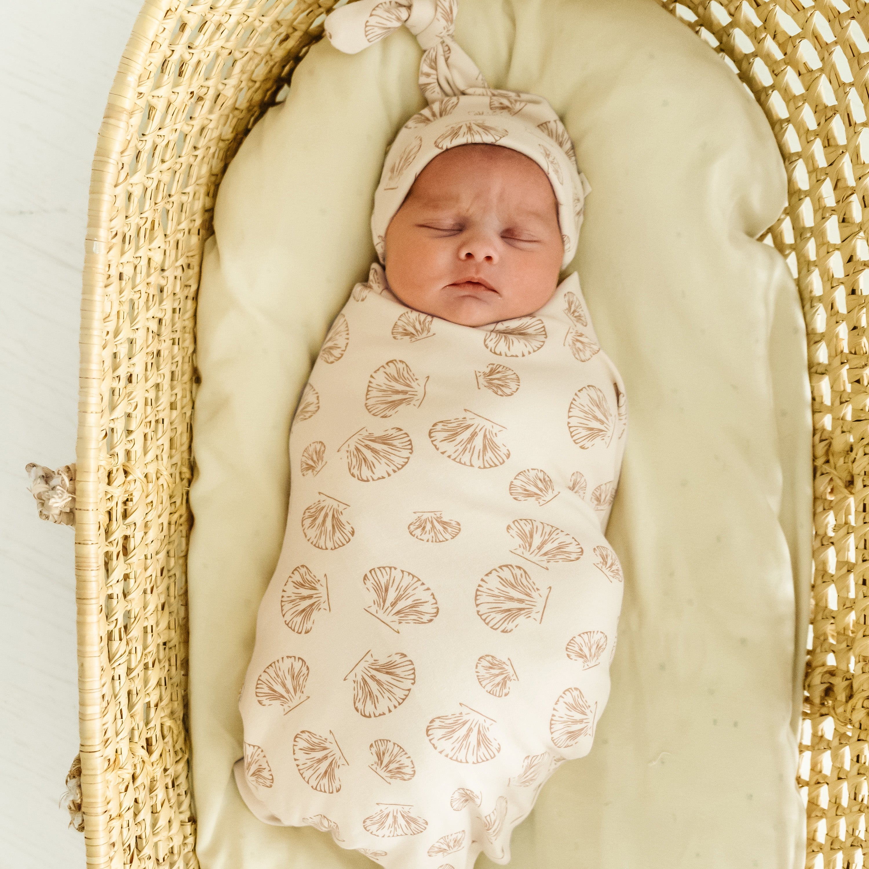 A newborn baby swaddled in a Makemake Organics cream-colored Organic Swaddle Blanket with a Seashells pattern, sleeping in a wicker bassinet on a light green cushion. The baby wears a matching headband.