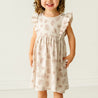 A smiling toddler with curly hair, wearing a Makemake Organics Organic Flutter Dress in Seashells pattern, stands in front of a plain, light-colored background.