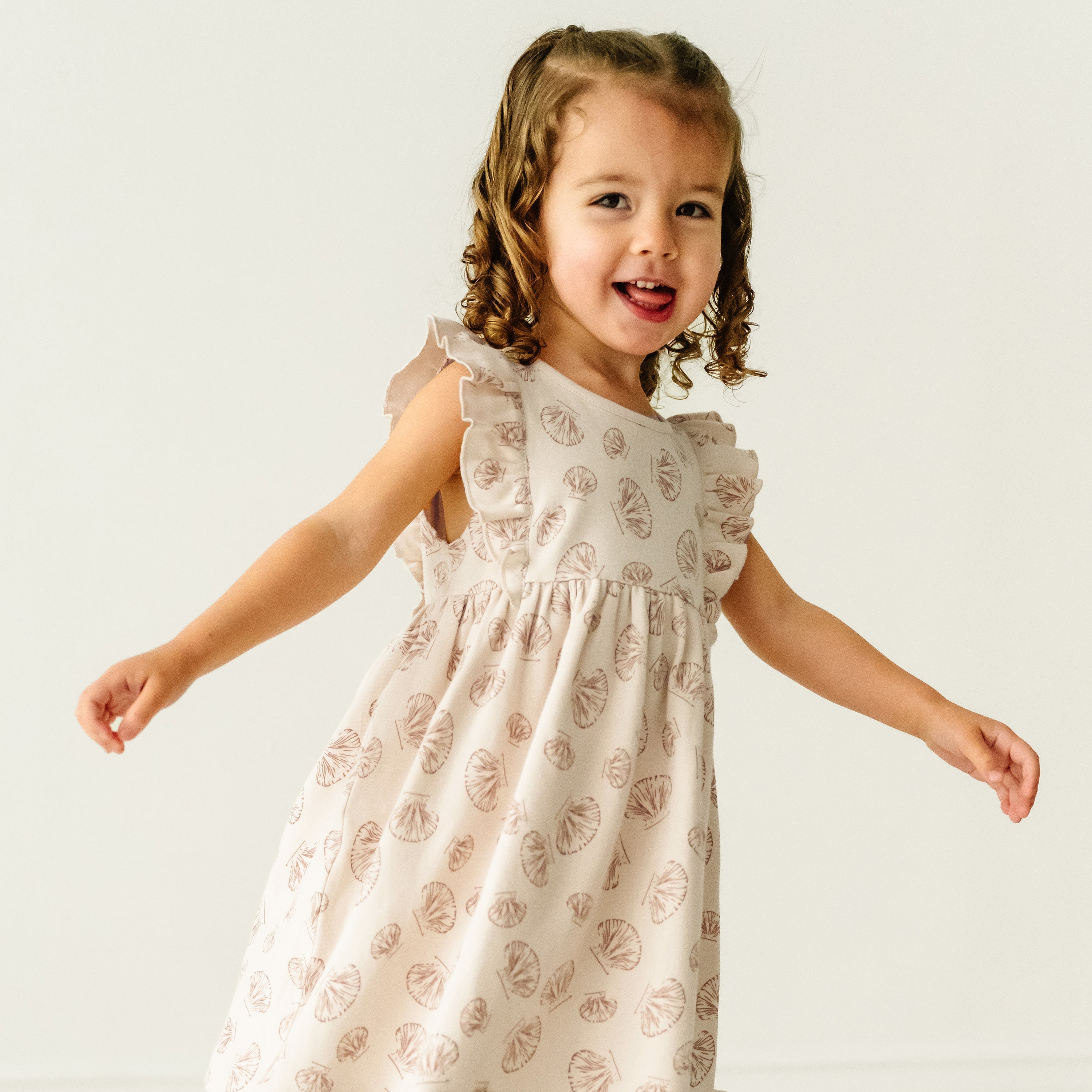 A joyful baby girl with curly hair, wearing a Makemake Organics Organic Flutter Dress - Seashells, smiles and stretches her arms out while standing against a plain light background.