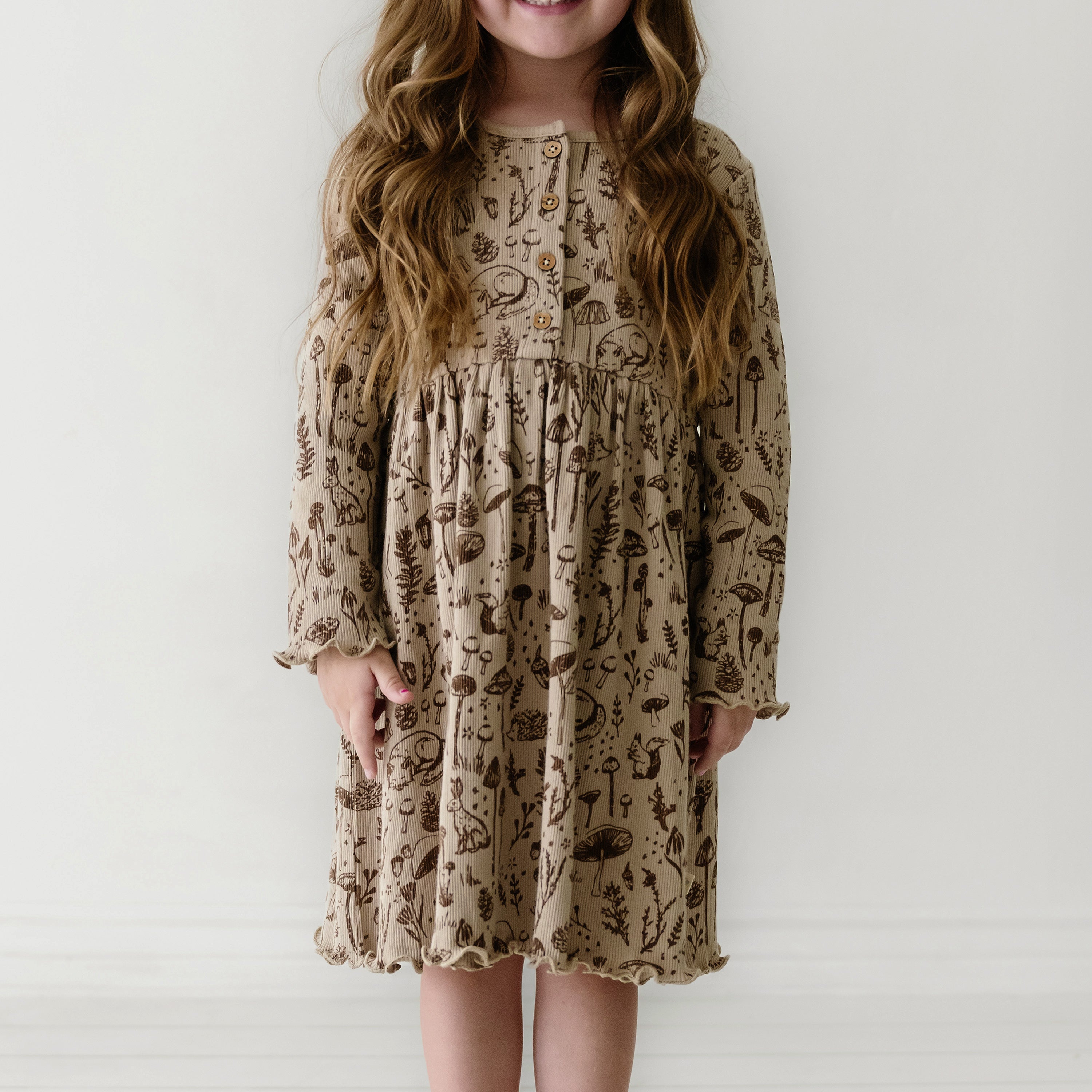 A young girl with long, wavy hair wearing an Organic Kids beige dress with botanical and animal prints, standing against a plain white background. She is smiling slightly while facing the camera.