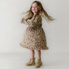 A joyful young girl with long, flowing hair spins in a Organic Long Sleeve Twirl Dress - Woodland from Organic Kids, wearing matching boots, against a plain white background.