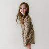 A young girl with long, wavy hair and a bright smile, wearing an Organic Kids Woodland organic long sleeve twirl dress, stands against a plain white background, looking over her shoulder.