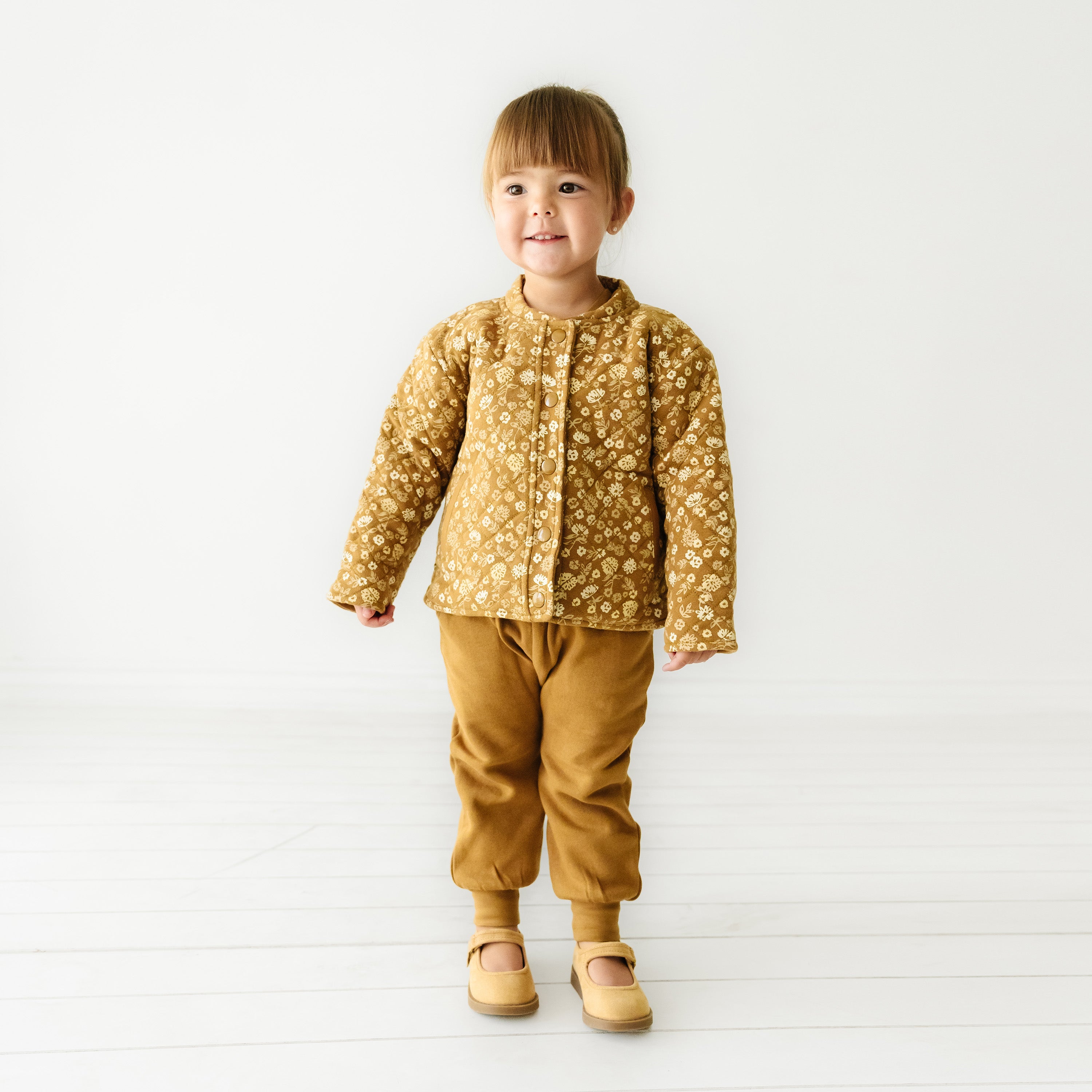 A toddler stands smiling in a studio, wearing an Organic Kids Wildflower organic merino wool buttoned jacket, taupe pants, and matching shoes against a plain white background.